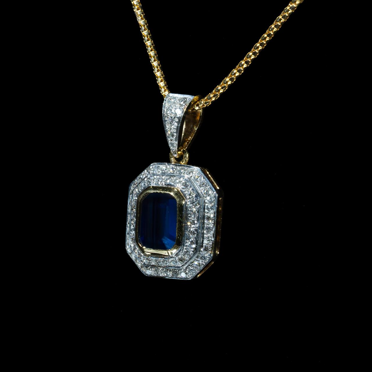 Elegant 18K yellow and white gold pendant necklace featuring a natural emerald-cut blue sapphire surrounded by a halo of brilliant natural diamonds suspended from a matching gold chain.